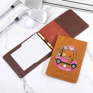 Könnte beinhalten: Ein brauner Golf-Scorekartenhalter aus Leder mit einem rosa Golfwagen-Design und dem Namen "Alexandra". Der Scorekartenhalter ist geöffnet und zeigt eine Scorekarte und einen Bleistift. Golfschläger und ein Golfball sind zu sehen.