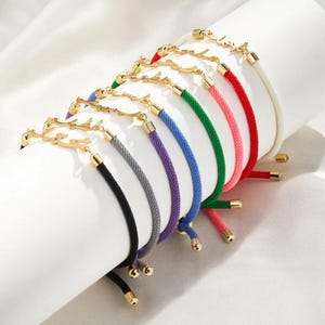 May include: A selection of colourful rope bracelets featuring gold-coloured antler charms. The bracelets are in various colours, including black, grey, purple, blue, green, pink, red, and white. Each bracelet has gold-coloured metal accents.
