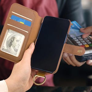 May include: Brown leather phone case with card slots and a cash pocket. The phone case is open, revealing a smartphone with a black screen. A person is holding the phone case, and another person is using a credit card.
