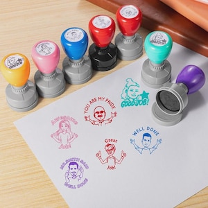 May include: A collection of seven self-inking stamps with colorful handles in yellow, pink, blue, red, and purple. Each stamp features a unique cartoon character illustration and text such as "Good Job," "Awesome Job," and "Well Done" printed on paper.