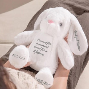 May include: A white plush bunny with long ears and a pink nose. The bunny has the text "Carried for a moment, loved for a lifetime" printed on its chest. The date "16.05.23" is printed on one foot, and "Baby Mattie" is printed on one ear.