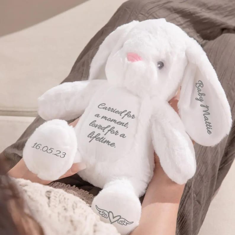 May include: A white plush bunny with long ears and a pink nose. The bunny has the text "Carried for a moment, loved for a lifetime" printed on its chest. The date "16.05.23" is printed on one foot, and "Baby Mattie" is printed on one ear.