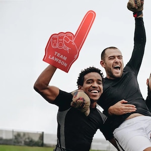 May include: Two people celebrating with a red foam finger that says "TEAM LAWSON". One person is holding the finger up in the air and the other person is holding onto their shoulders.