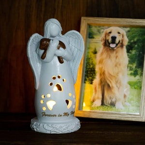 Op de afbeelding: Witte keramische engelfiguur die een kleine bruine hond vasthoudt. De engel heeft vleugels en een halo. De figuur heeft poot afdrukken die uit het lichaam zijn gesneden en de woorden "Forever in My Heart" op de basis.