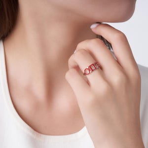 May include: A red ring with the word "ONE" in a minimalist design. The ring features a circle, a rectangle, and another rectangle, forming the letters. The ring is worn on a finger.
