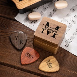 May include: Wooden guitar pick set with a personalized wooden box. The box is engraved with the name "Madison" and the initial "M". Three guitar picks are engraved with initials and dates. The picks are various shades of brown.
