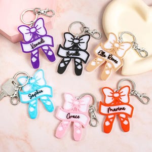 Può includere: Una collezione di portachiavi a tema balletto in vari colori, tra cui viola, nero, beige, blu, rosa e arancione. Ogni portachiavi presenta un fiocco, scarpette da ballo e un nome su uno striscione. I portachiavi hanno portachiavi e chiusure color argento.