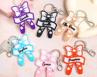 Puede incluir: Una colección de llaveros con temática de ballet en varios colores, incluyendo morado, negro, beige, azul, rosa y naranja. Cada llavero presenta un lazo, zapatillas de ballet y un nombre en una pancarta. Los llaveros tienen llaveros y cierres plateados.