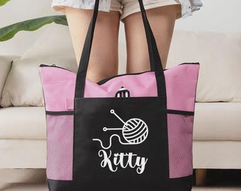 May include: A pink and black tote bag with a white graphic of yarn and knitting needles, and the word "Kitty" in script. The bag has black handles and mesh side pockets. The bag is held by a person.