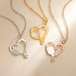 May include: Three heart-shaped pendant necklaces. One is silver with a badminton racket design and the name "Bonnie". The second is gold with a ping pong paddle design and the name "Helen". The third is rose gold with a pickleball paddle design and the name "Charlotte".