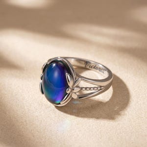 May include: Silver ring featuring an oval, colour-changing stone that transitions from blue to purple. Floral details decorate the sides, and the name "Victoria" is engraved on the band. The ring is displayed on a neutral background.