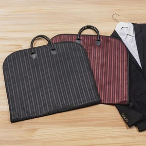 Puede incluir: Tres fundas para prendas con asas. Una es negra con rayas verticales, otra es burdeos con rayas verticales y la tercera es negra. Una chaqueta de traje negra y una camisa blanca est&aacute;n en una percha en el fondo.