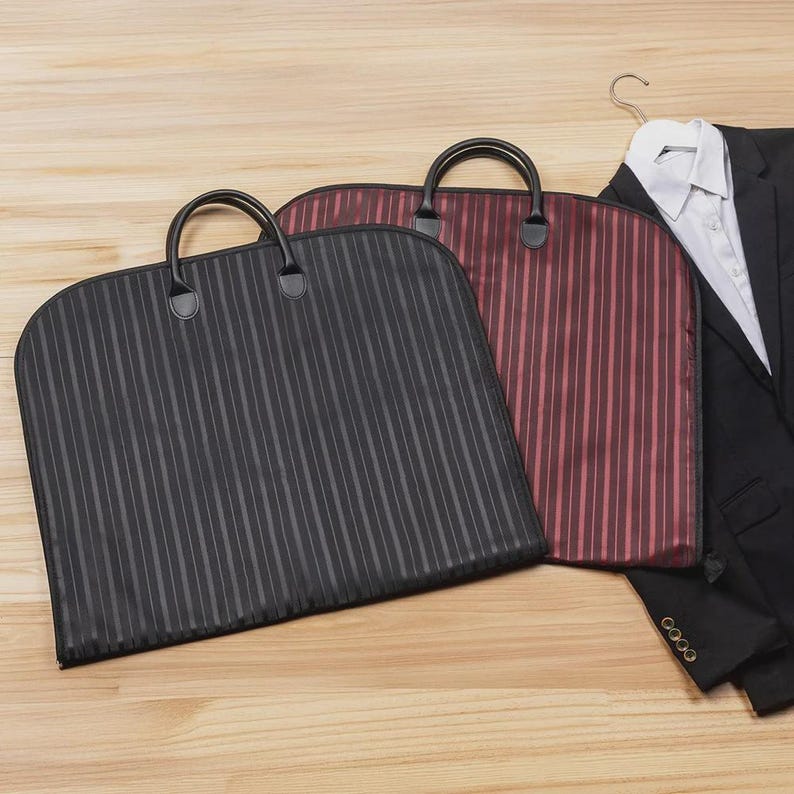 Puede incluir: Tres fundas para prendas con asas. Una es negra con rayas verticales, otra es burdeos con rayas verticales y la tercera es negra. Una chaqueta de traje negra y una camisa blanca est&aacute;n en una percha en el fondo.