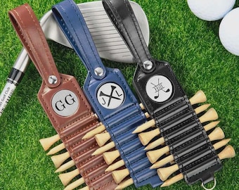 May include: Three leather golf tee holders in brown, blue, and black, each with a different initial design. The holders are filled with wooden golf tees and are on a bed of green grass. A golf club and golf balls are in the background.