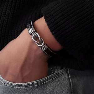 Könnte beinhalten: Ein schwarzes Lederarmband mit einem silbernen Knotenschließe.
