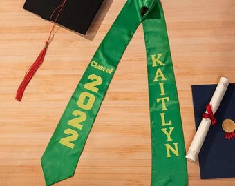Puede incluir: Banda de graduación verde con letras doradas. La banda dice "Class of 2022" en un lado y "Kaitlyn" en el otro.
