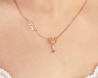 May include: A rose gold necklace with a delicate chain. The pendant features a rose with a small purple gemstone and the name "Ruby" in cursive script. The necklace is worn around the neck.