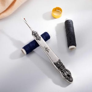 A metal tool with a sharp tip and a hook that can cut threads and seams. The tool can be customized with a name engraved on the handle. It is a vintage style sewing gift for tailors, seamstresses, or fashion designers.