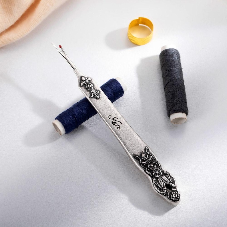 A metal tool with a sharp tip and a hook that can cut threads and seams. The tool can be customized with a name engraved on the handle. It is a vintage style sewing gift for tailors, seamstresses, or fashion designers.
