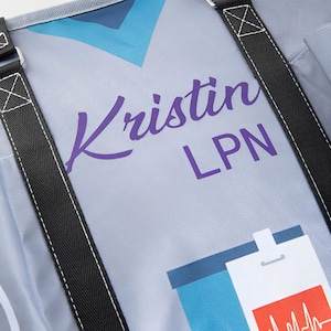 May include: A gray canvas tote bag with black straps. The bag has a purple inscription that says "Kristin LPN" and a blue and white graphic of a stethoscope.