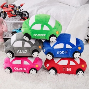 May include: A stack of plush toy cars in various colors: green, gray, blue, pink, and red. Each car has a white roof and black wheels. Names are printed on the side of each car: "COOPER", "ALEX", "EDDIE", "OLIVIA", and "TIM".
