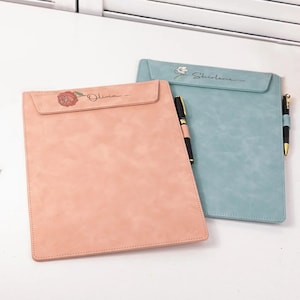 May include: Two leatherette clipboards, one pink and one teal, with matching pens. The pink clipboard features a rose and the name "Olivia". The teal clipboard has a floral design and the name "Shirleyne".