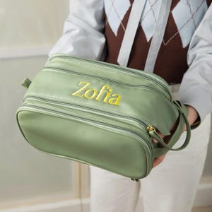 Può includere: Una borsa da viaggio verde salvia con più scomparti con cerniera. La parola "Zofia" è ricamata con filo giallo sul davanti. La borsa ha un manico laterale e cerniere color oro.