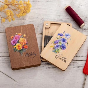 Puede incluir: Dos cajas de madera con diseños florales y nombres personalizados, "Ashley" y "Ella". Cada caja contiene un juego de agujas.