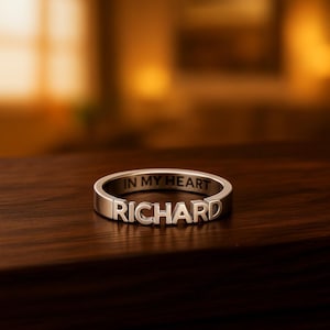 Könnte beinhalten: Ein silberner Ring mit dem Namen "RICHARD" in erhabenen Buchstaben. Die Worte "IN MY HEART" sind auf der Oberseite des Rings eingraviert. Der Ring liegt auf einer dunklen Holzoberfläche.