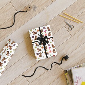 Puede incluir: Papel de regalo blanco con un patrón repetido de perros marrones y flores de colores. Una cinta negra está atada alrededor de la caja de regalo.