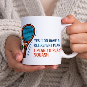 Könnte beinhalten: Weiße Keramiktasse mit einer Squashschläger-Grafik und dem Text "YES, I DO HAVE A RETIREMENT PLAN I PLAN TO PLAY SQUASH" in Blau und Orange. Die Tasse hat einen gebogenen Henkel und wird von einer Person gehalten.