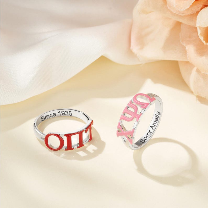May include: Two silver rings with Greek letters in red and pink enamel. One ring has "Since 1935" engraved, the other has "Soror Amelia" engraved. The rings are on a cream-colored surface.
