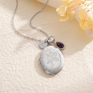 May include: A silver-colored locket necklace with a floral design, a small initial charm, and a purple gemstone pendant. The oval locket is engraved with a flower and leaf pattern. The necklace is on a delicate chain.