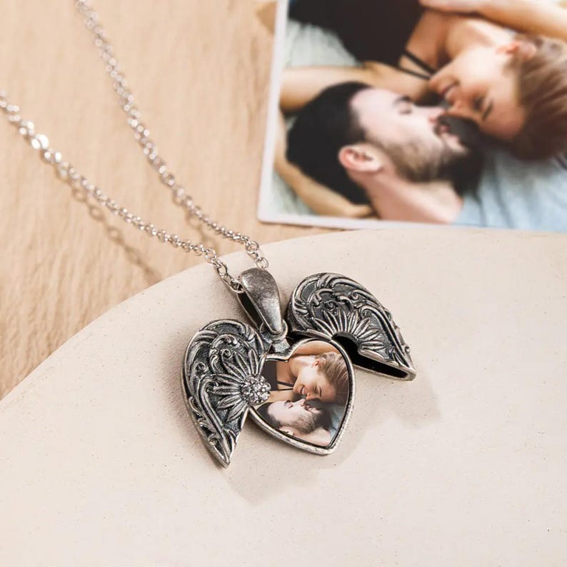 May include: Silver locket necklace with a heart-shaped opening that holds a photo of a couple. The locket is decorated with intricate details and has a small crystal on the front.