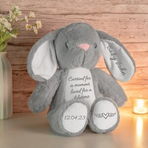 May include: A gray plush bunny with a pink nose and inner ears. The bunny has the text "Baby John" on one ear and "Carried for a moment, loved for a lifetime" on its chest. The date "12.04.23" and a heart with wings are on the foot.