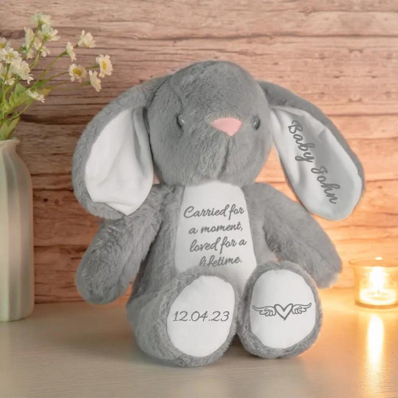 May include: A gray plush bunny with a pink nose and inner ears. The bunny has the text "Baby John" on one ear and "Carried for a moment, loved for a lifetime" on its chest. The date "12.04.23" and a heart with wings are on the foot.