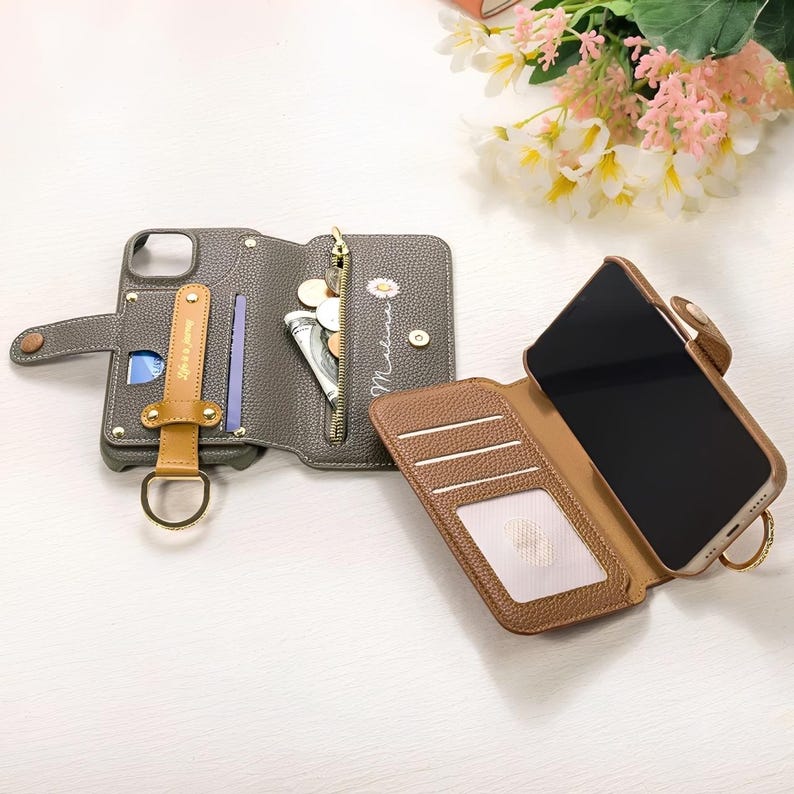 May include: Gray and brown leather phone case with a wallet design. The case has a zipper pocket, card slots, and a wrist strap. A daisy and the name "Melina" are printed on the case. The phone case is open, revealing a black screen.