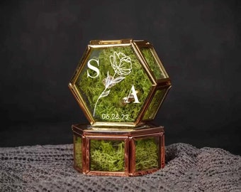 May include: A hexagonal glass ring box with a gold frame and a lid. The box is filled with green moss and has a white floral design with the initials "S" and "A" and the date "06.26.22" engraved on the lid.