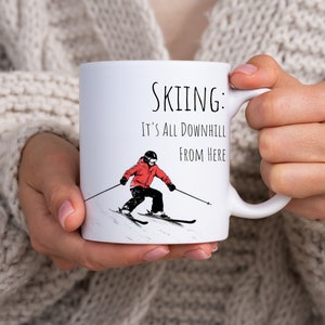 May include: White ceramic mug with a black and white illustration of a skier on a snowy mountain. The text "SKIING: IT'S ALL DOWNHILL FROM HERE" is printed on the mug.