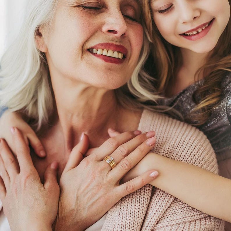 Pu&ograve; includere: Primo piano di due persone che si abbracciano, una pi&ugrave; anziana con i capelli grigi e l'altra pi&ugrave; giovane con i capelli biondi. La persona anziana indossa un anello d'oro con una pietra rettangolare. La persona anziana indossa un maglione rosa chiaro.