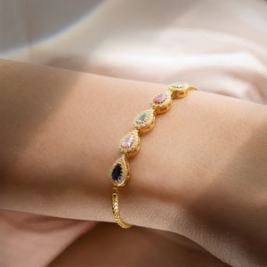 Op de afbeelding: Een gouden armband met een ketting en zes druppelvormige stenen die zijn gezet in een halo van kleinere stenen. De stenen zijn blauw, groen, roze, rood en helder.
