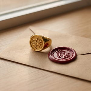 May include: A gold signet ring featuring a raised lion emblem on its circular face, positioned next to a round, deep red wax seal that displays an identical lion motif, both resting on a light brown paper envelope surface.
