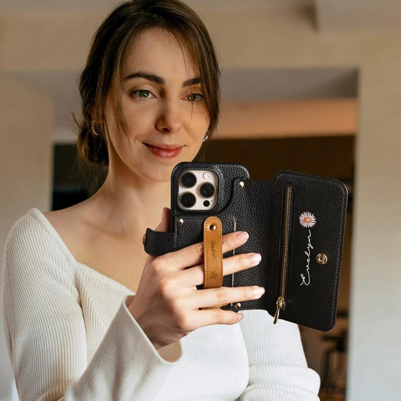 May include: Black leather phone case with a wallet feature. The case has a gold zipper, a daisy graphic, and the name "Evelyn" in cursive. A tan leather hand strap is attached to the phone case.
