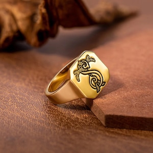 Könnte beinhalten: Ein goldfarbener Ring mit einem geometrischen Design auf der Oberfläche. Der Ring hat eine rechteckige Oberfläche mit einem detaillierten schwarzen Design. Der Ring ist vor einem braunen Hintergrund platziert.