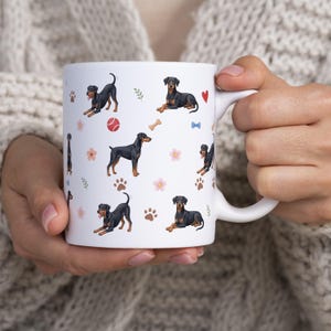 May include: A white ceramic mug featuring a repeating pattern of black and tan Doberman dogs in various poses, interspersed with small red hearts, pink flowers, brown paw prints, a red ball, a tan bone, and a small blue bow.