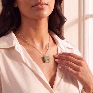 May include: A gold chain necklace featuring a small, rectangular, sage green book-shaped pendant. The pendant displays a miniature illustration of a peach with leaves and the text "The Orchard in Winter Hill" printed on the front cover in gold lettering.