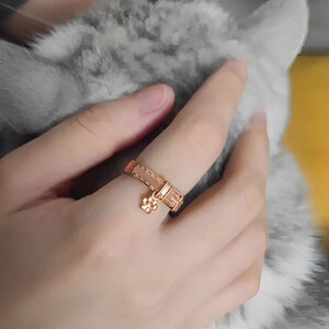 May include: A rose gold ring with a paw print charm. The ring is designed to look like a dog collar.