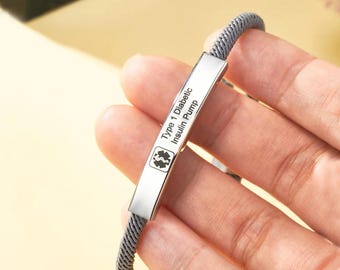 May include: A silver medical ID bracelet with a grey woven band. The rectangular plate is engraved with "Type 1 Diabetic Insulin Pump" and a medical symbol. The bracelet is designed for medical alert purposes.