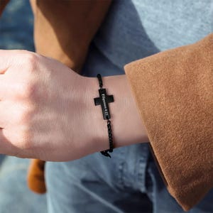 May include: Black bracelet featuring a cross pendant with the inscription "Philippians 4:13". The bracelet has a chain-like design and is worn on a wrist. The cross is a prominent, dark shape against the skin.