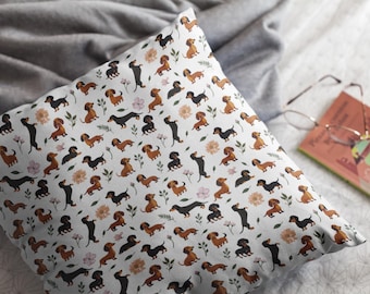 May include: Square white pillow with a repeating pattern of dachshund dogs in various poses, interspersed with floral and leaf elements. The dogs are in shades of brown and black. A book and glasses are in the background.